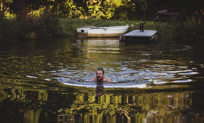 A man swims in the foreground.  the scene is of a pond with a jetty in the distance on which stands a dog with a rowing boat just seen.